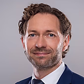 Porträt eines Mannes mit lockigem Haar, der in einem Anzug mit Krawatte in die Kamera lächelt. Heller Hintergrund.