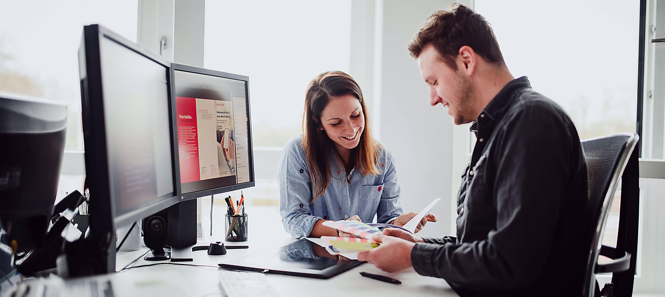 Ein Mann und eine Frau in einem Büro besprechen Dokumente, während sie an einem Tisch mit Computer und Schreibutensilien sitzen.