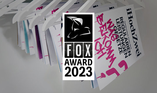 Badge für die Auszeichnung Fox Award 2023 in silber auf den prämierten Zeitschriften, welche im Hintergrund auf Kleiderbügeln hängen