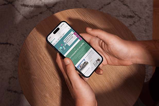 Eine Hand hält ein Smartphone, das eine Webseite mit Informationen und Grafiken anzeigt, auf einem Holztisch platziert.