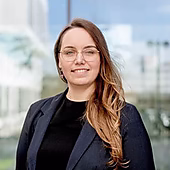 Eine lächelnde Frau mit langen Haaren in einem Blazer steht vor einem modernen Gebäude mit großen Fenstern.